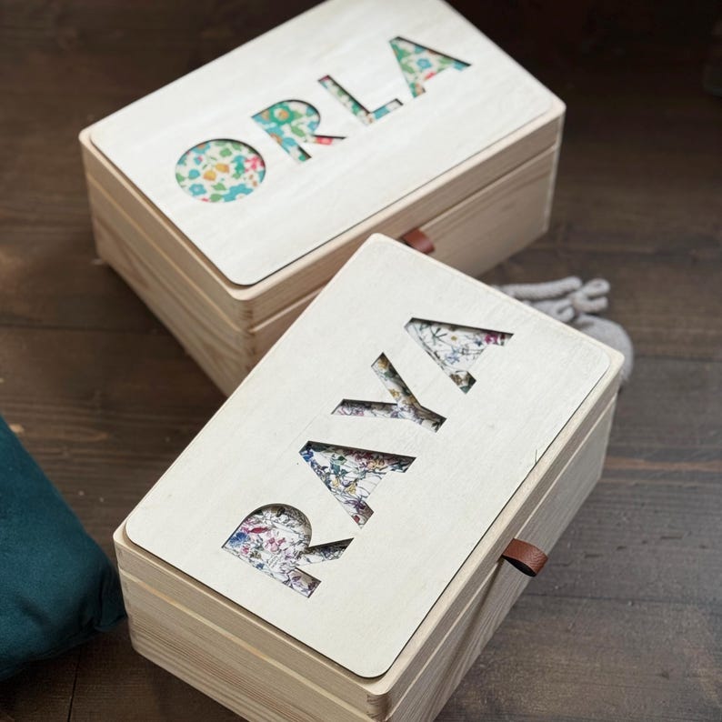 K&ouml;nnte beinhalten: Zwei Holzkisten mit ausgeschnittenen Buchstaben, die "ORLA" und "RAYA" auf den Deckeln bilden. Die ausgeschnittenen Buchstaben zeigen einen floralen Stoff. Jede Box hat eine kleine Lederlasche an der Seite.