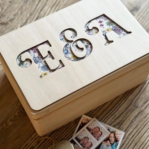 May include: A wooden box with a floral pattern cut out to reveal the letters "E & A". The box is closed and sitting on a wooden surface.