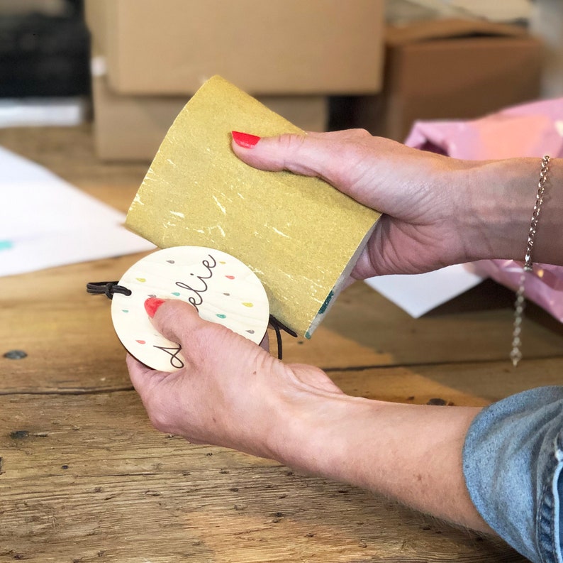May include: A round wooden tag with the name "Alie" written in cursive. The tag has a colorful confetti design and a brown string attached to it. A person is holding the tag in their hand.