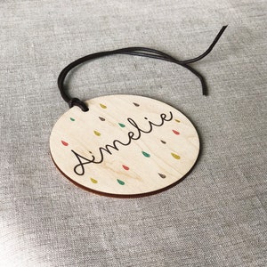 May include: A round wooden ornament with a brown leather cord. The ornament has a white background with colorful raindrops and the name "Amelie" written in black cursive.