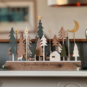 Pode incluir: Calend&aacute;rio do advento de Natal em madeira com uma cena de inverno com &aacute;rvores, uma casa, veados e uma lua crescente. A base &eacute; gravada com as palavras "all is calm, all is bright". As &aacute;rvores s&atilde;o em v&aacute;rios tons de verde, castanho e branco.