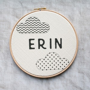 May include: A white canvas with a wooden frame, featuring two black and white cloud designs and the name "ERIN" in black lettering.