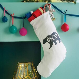 Può includere: Calza di Natale bianca con una sagoma di orso nero e il nome "Oscar" stampato su di essa. La calza è appesa a un camino con una ghirlanda di nastro blu e bianco e decorazioni.