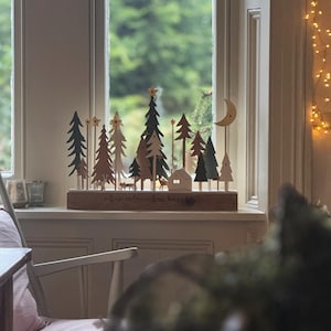 Pode incluir: Uma cena de Natal em madeira com &aacute;rvores, uma casa, veados e uma lua crescente. A base diz "all is calm, all is bright". A cena est&aacute; situada numa janela com um fundo verde desfocado e luzes de fadas.