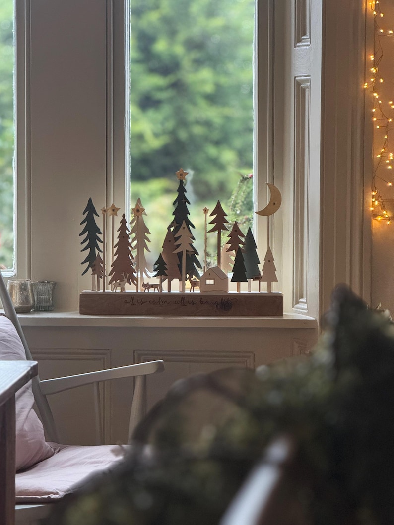 Pode incluir: Uma cena de Natal em madeira com &aacute;rvores, uma casa, veados e uma lua crescente. A base diz "all is calm, all is bright". A cena est&aacute; situada numa janela com um fundo verde desfocado e luzes de fadas.