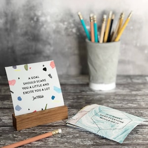 Puede incluir: Una tarjeta de cita motivacional con un fondo abstracto colorido. La cita dice "Una meta debe asustarte un poco y emocionarte mucho." - Joe Vitale. La tarjeta se muestra en un soporte de madera.