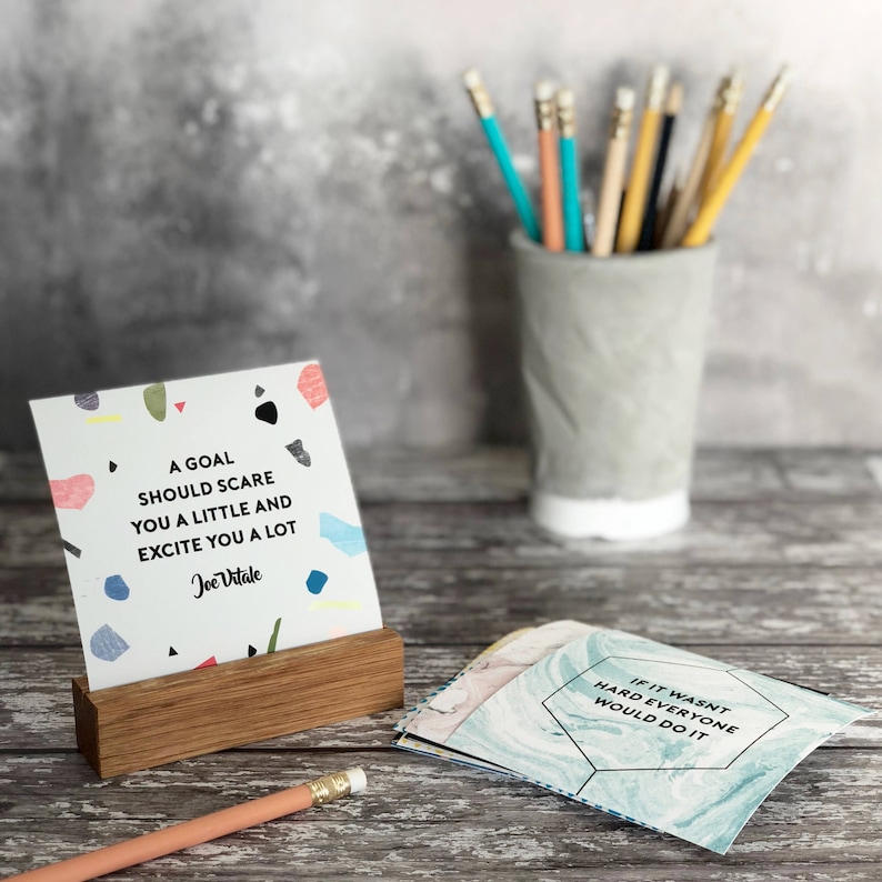 Puede incluir: Una tarjeta de cita motivacional con un fondo abstracto colorido. La cita dice "Una meta debe asustarte un poco y emocionarte mucho." - Joe Vitale. La tarjeta se muestra en un soporte de madera.