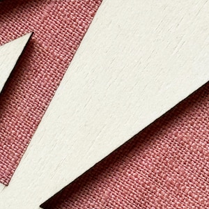 K&ouml;nnte beinhalten: Nahaufnahme eines Holzausschnitts mit der Zahl 17. Die Zahl ist wei&szlig;, mit einem strukturierten, r&ouml;tlich-rosa Stoff, der durch den negativen Raum sichtbar ist. Das Holz hat eine helle, nat&uuml;rliche Oberfl&auml;che.