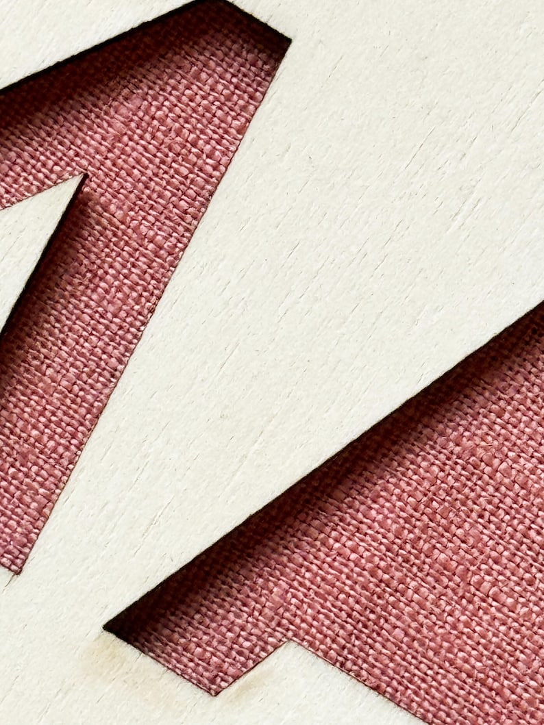 K&ouml;nnte beinhalten: Nahaufnahme eines Holzausschnitts mit der Zahl 17. Die Zahl ist wei&szlig;, mit einem strukturierten, r&ouml;tlich-rosa Stoff, der durch den negativen Raum sichtbar ist. Das Holz hat eine helle, nat&uuml;rliche Oberfl&auml;che.