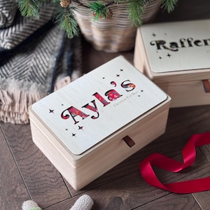 Könnte beinhalten: Eine rechteckige, helle Holzbox mit einem Deckel, der den ausgeschnittenen Namen Ayla's und den Text Christmas Eve Box sowie kleine Sternmotive zeigt. Das Innere des Ausschnitts offenbart ein rotes Blumenmuster. Eine braune Lederlasche ist angebracht.