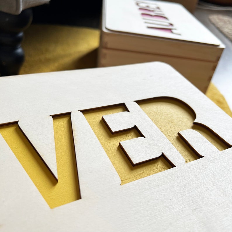 May include: Close-up of a wooden sign with the word "VER" cut out, revealing a yellow background. Another wooden box with the name "AUBREY" is visible in the background. The sign and box are made of light-colored wood.