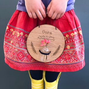 Op de afbeelding: Een ronde houten plaat met de naam "Cora" en de tekst "expert finder of teeny tiny treasures for mummy". Een kleine glazen fles met roze bloemen is bevestigd. Het bord wordt opgehangen met een zwart touwtje.