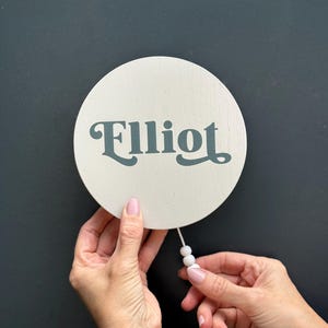 Könnte beinhalten: Ein runder, cremefarbener Holzschild mit dem Namen "Elliot" in türkisfarbener Schrift. Das Schild hat eine Schnur mit weißen Perlen. Der Hintergrund ist dunkelgrau.