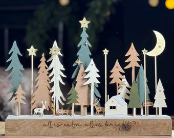 May include: Wooden Christmas advent calendar with a variety of trees, stars, a moon, and a small house. The base is engraved with "all is calm, all is bright." The trees are in shades of green, white, and brown. Includes small deer figurines.