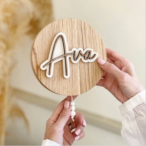 May include: A round wooden sign with the name "Ava" written in white script on a brown background. A string of white beads hangs from the bottom of the sign.