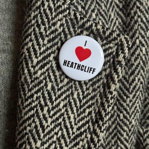 Könnte beinhalten: Ein weißer Ansteckbutton mit dem Text "I HEART HEATHCLIFF" in Schwarz und einem roten Herz. Der Button ist an einem schwarz-weiß karierten Stoff befestigt.