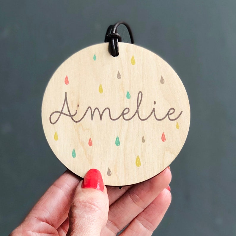 May include: A wooden oval ornament with a brown leather cord. The ornament is decorated with a colorful raindrop pattern and the name "Amelie" written in cursive.