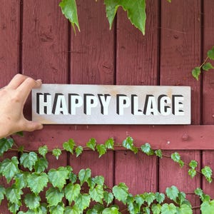Könnte beinhalten: Ein rechteckiges Holzschild mit heller Oberfläche, das den Schriftzug HAPPY PLACE in fetten, weißen Blockbuchstaben mit schwarzem Schlagschatten zeigt, wodurch ein dreidimensionaler Effekt auf der Oberfläche des Schildes entsteht.