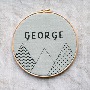 May include: A light gray canvas with the name "GEORGE" in black lettering, surrounded by three black and white geometric mountain designs. The canvas is mounted in a wooden hoop.