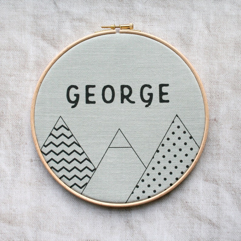 May include: A light gray canvas with the name "GEORGE" in black lettering, surrounded by three black and white geometric mountain designs. The canvas is mounted in a wooden hoop.