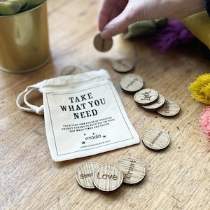 May include: A small canvas bag with a drawstring closure contains wooden tokens with positive words such as "Love", "Trust", "Strength", "Confidence", "Energy", and "Luck". The bag has the text "Take What You Need" printed on it.