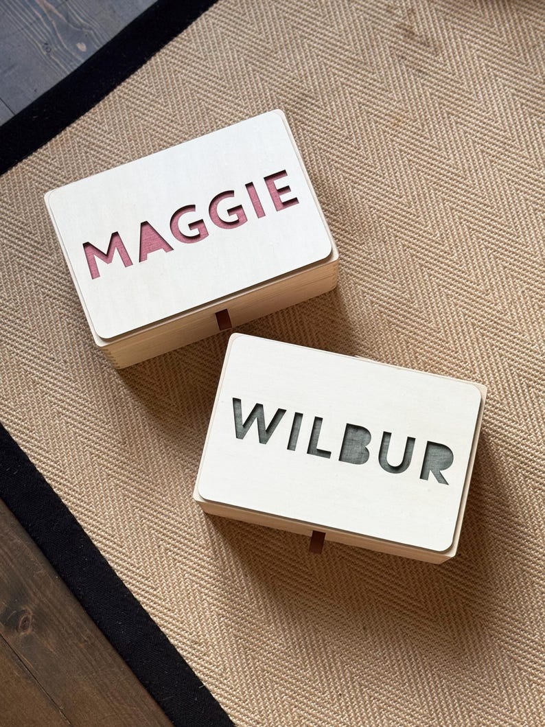 K&ouml;nnte beinhalten: Zwei Holzkisten mit ausgeschnittenen Namen, "MAGGIE" und "WILBUR", werden gezeigt. Die Namen sind durch die ausgeschnittenen Buchstaben sichtbar. Die Innenseite der "MAGGIE"-Box ist rosa und die "WILBUR"-Box ist gr&uuml;n. Die Boxen stehen auf einer beigefarbenen Fischgr&auml;tmuster-Oberfl&auml;che.