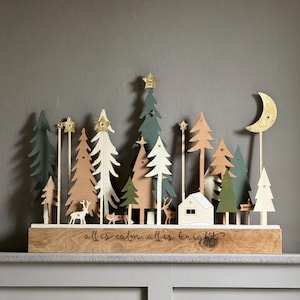 Pode incluir: Calend&aacute;rio do advento de Natal em madeira com uma cena de floresta com &aacute;rvores, veados e uma casa. As &aacute;rvores s&atilde;o em v&aacute;rios tons de verde, branco e madeira natural, com estrelas douradas. A base diz "all is calm, all is bright?"