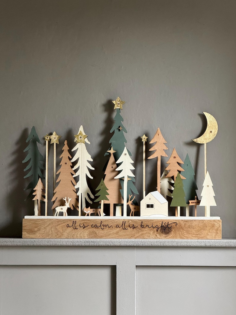 Pode incluir: Calend&aacute;rio do advento de Natal em madeira com uma cena de floresta com &aacute;rvores, veados e uma casa. As &aacute;rvores s&atilde;o em v&aacute;rios tons de verde, branco e madeira natural, com estrelas douradas. A base diz "all is calm, all is bright?"