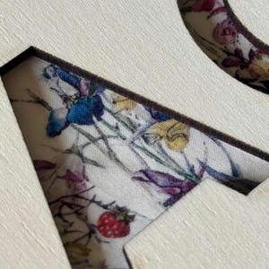 K&ouml;nnte beinhalten: Nahaufnahme eines Holzhandwerks mit einem floralen Stoffeinsatz. Der Stoff weist ein Muster aus bunten Blumen auf, darunter blaue, violette und rote Bl&uuml;ten, vor einem cremefarbenen Hintergrund. Der Holzrahmen hat eine nat&uuml;rliche, helle Farbe.