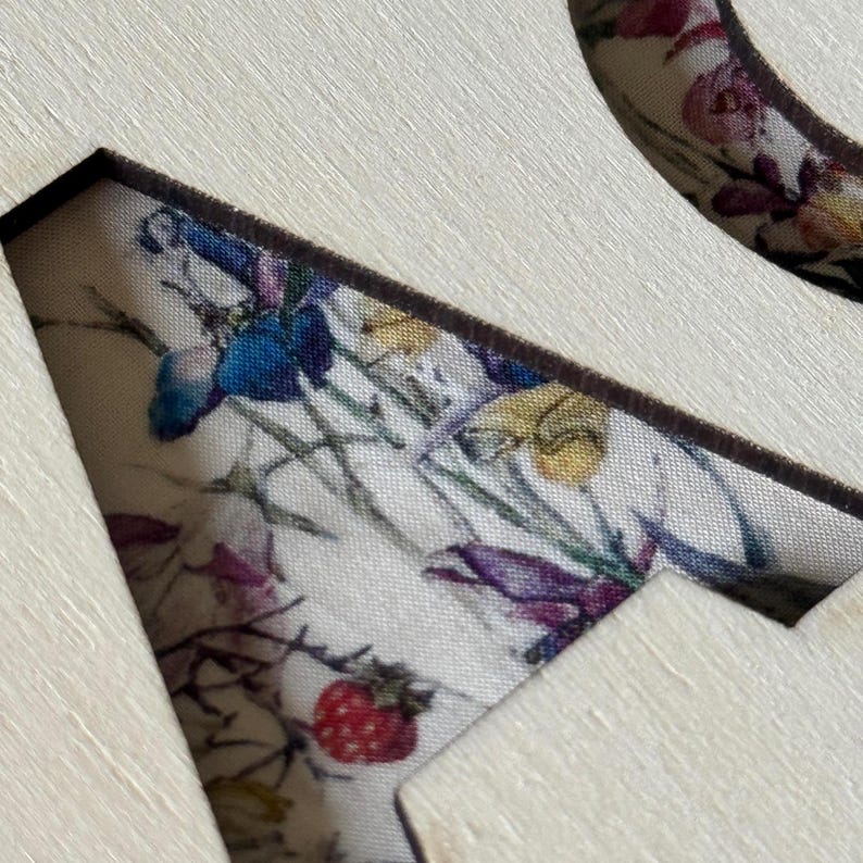 K&ouml;nnte beinhalten: Nahaufnahme eines Holzhandwerks mit einem floralen Stoffeinsatz. Der Stoff weist ein Muster aus bunten Blumen auf, darunter blaue, violette und rote Bl&uuml;ten, vor einem cremefarbenen Hintergrund. Der Holzrahmen hat eine nat&uuml;rliche, helle Farbe.
