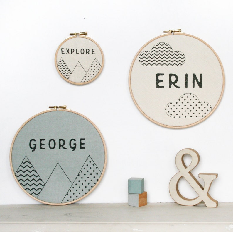 May include: Three round embroidery hoops with different designs. The top hoop has a white background with black clouds and the name "ERIN" in black text. The middle hoop has a light blue background with black mountains and the name "GEORGE" in black text. The bottom hoop has a white background with black mountains and the text "EXPLORE" in black text. There is a wooden ampersand symbol and two small wooden blocks.