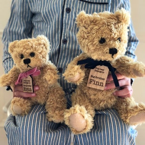 May include: Two plush teddy bears with personalized wooden tags. The larger bear has a black ribbon and a tag that reads "This bear belongs to Big Brother Finn". The smaller bear has a pink ribbon and a tag that reads "This bear belongs to Baby Sister Erin".