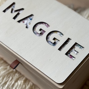 K&ouml;nnte beinhalten: Eine hellfarbene Holzkiste mit dem Namen "MAGGIE" aus dem Deckel ausgeschnitten. Die ausgeschnittenen Buchstaben zeigen darunter ein Blumenmuster. Ein kleines braunes Lederband ist an der Seite der Box befestigt.