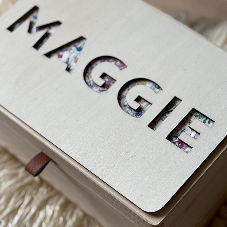 K&ouml;nnte beinhalten: Eine hellfarbene Holzkiste mit dem Namen "MAGGIE" aus dem Deckel ausgeschnitten. Die ausgeschnittenen Buchstaben zeigen darunter ein Blumenmuster. Ein kleines braunes Lederband ist an der Seite der Box befestigt.