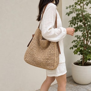 Peut inclure: Un sac fourre-tout en paille tressée avec des lanières en cuir marron. Le sac a un grand compartiment principal et une petite poche. Le sac est de couleur beige et marron. Le sac est porté à l'épaule.