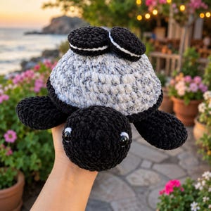 Peut inclure: Une peluche tortue crochetée à la main avec une tête et des nageoires noires, dotée d'une carapace texturée mouchetée de blanc et de noir. Deux petites décorations rondes en forme de biscuit sandwich noir et blanc sont fixées sur le dessus.