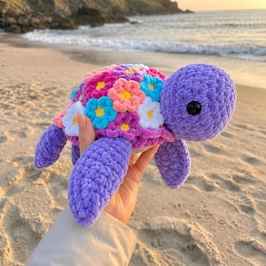 Peut inclure: Une tortue en peluche faite de fil violet avec une carapace décorée de nombreuses petites fleurs crochetées multicolores dans des tons de rose, bleu, blanc et jaune. La tortue a une tête ronde avec des yeux en plastique noir et quatre nageoires.
