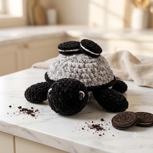 May include: A crocheted turtle plush with a black head and flippers, and a speckled gray and white shell. Two small sandwich cookies are attached to the top of the shell, and two additional sandwich cookies sit nearby with scattered crumbs.