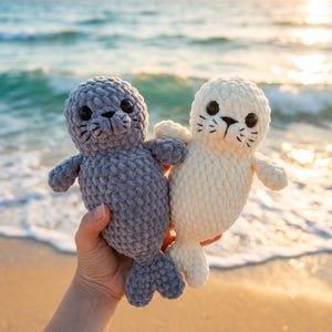 Peut inclure: Deux jouets phoques en peluche faits de fil épais et doux. Un phoque est gris foncé et l'autre est blanc crème. Tous deux présentent des yeux ronds en plastique noir, des nez noirs brodés et des moustaches cousues avec des corps arrondis.