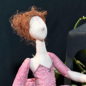 Puede incluir: Muñeca hecha a mano vestida de bailarina, con un vestido rosa con estampado floral y medias a juego. La muñeca tiene el pelo rojo y está sentada sobre un cojín de terciopelo dorado. Una linterna negra y hojas verdes en el fondo.