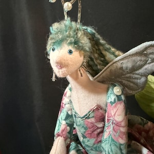 Puede incluir: Una muñeca de hada hecha a mano con una cara rosa suave, ojos azules y cabello turquesa. La muñeca viste un vestido con estampado floral con alas plateadas y antenas de cuentas. Los brazos de la muñeca están adornados con botones blancos.