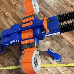 May include: A blue and orange toy blaster with a large rotating drum and two black barrels. The blaster has the word "ELITE" printed on it. A yellow measuring tape is next to the blaster.