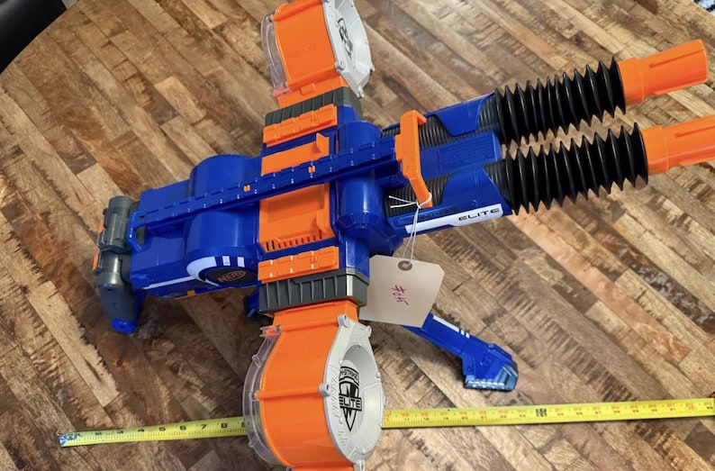 May include: A blue and orange toy blaster with a large rotating drum and two black barrels. The blaster has the word "ELITE" printed on it. A yellow measuring tape is next to the blaster.