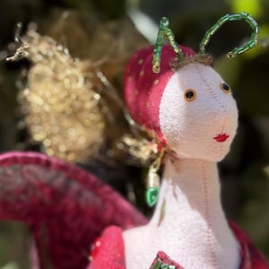 Op de afbeelding: Een handgemaakte stoffen pop met een grillig ontwerp. De pop heeft een wit gezicht, rode lippen en zwarte ogen. Het heeft een rood-witte gestippelde hoofdbedekking, groene kralenantennes en rood gevederde vleugels. De pop is versierd met groene kralenaccenten en een rode knoop.