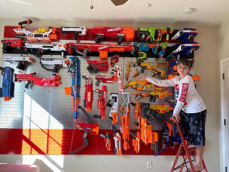 May include: A display of toy blasters in a range of colours, including orange, red, white, and blue, mounted on a wall. The toy guns are of different sizes and designs. A person is standing on a red ladder in front of the wall, pointing at the display.