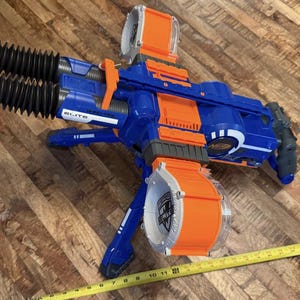 May include: A blue and orange toy gun with two orange barrels and a large orange drum magazine. The toy gun has the word "ELITE" printed on it. A yellow measuring tape is in the foreground.