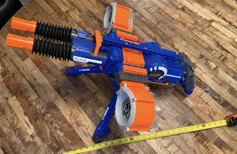 May include: A blue and orange toy gun with two orange barrels and a large orange drum magazine. The toy gun has the word "ELITE" printed on it. A yellow measuring tape is in the foreground.