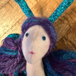 May include: A handmade doll with a white face, blue eyes, and a small red mouth. The doll has purple and blue yarn hair and teal fabric antennae. The doll is a whimsical, handcrafted item.