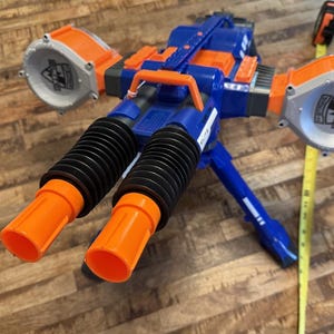 May include: A blue and orange toy gun with two orange barrels and two large orange and grey circular attachments. The toy gun has a black accordion-style section and a blue tripod base. A yellow measuring tape is next to the toy gun.