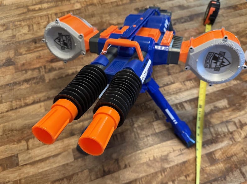 May include: A blue and orange toy gun with two orange barrels and two large orange and grey circular attachments. The toy gun has a black accordion-style section and a blue tripod base. A yellow measuring tape is next to the toy gun.